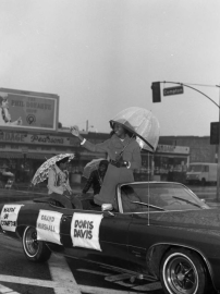 Doris Davis riding in convertible in rain as parade Grand Marshall.