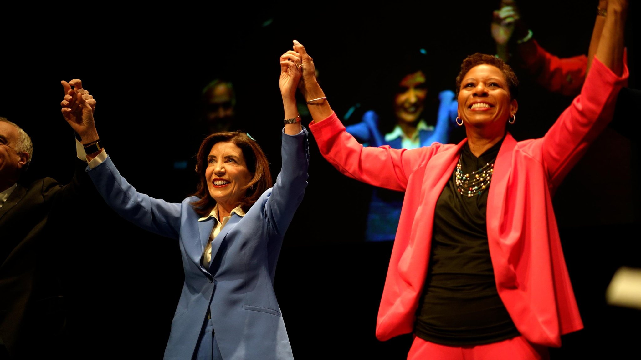 hochul and adams holding hands up to celebrate on stage
