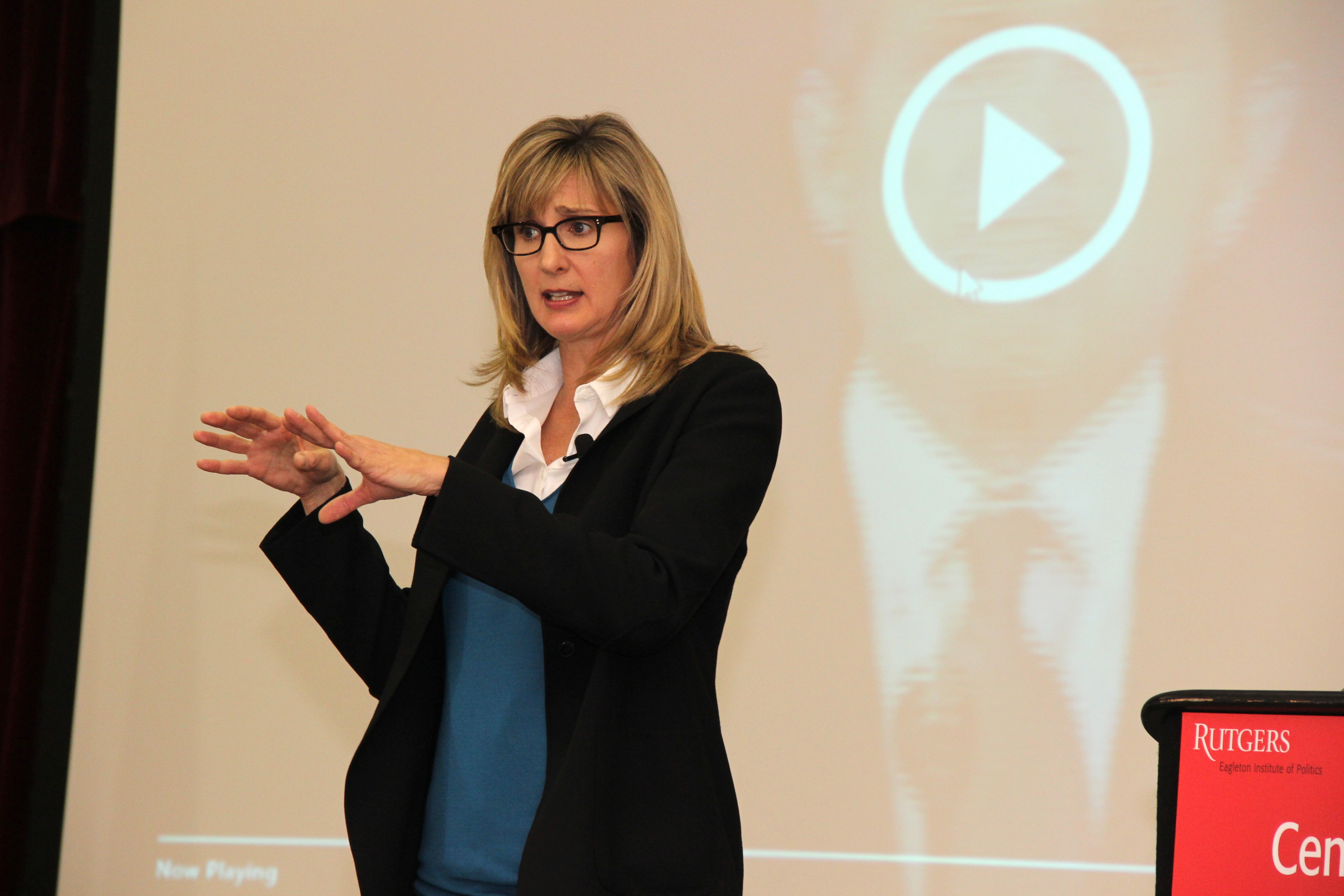 Woman with blonde hair and glasses standing on stage in front of projector screen, wearing a white shirt, blue sweater, and black blazer. 