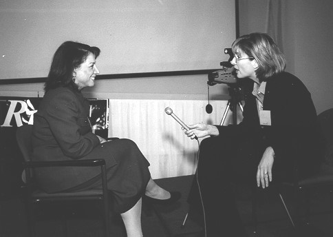 Two women sit in facing chairs, with one woman holding microphone and interviewing the other