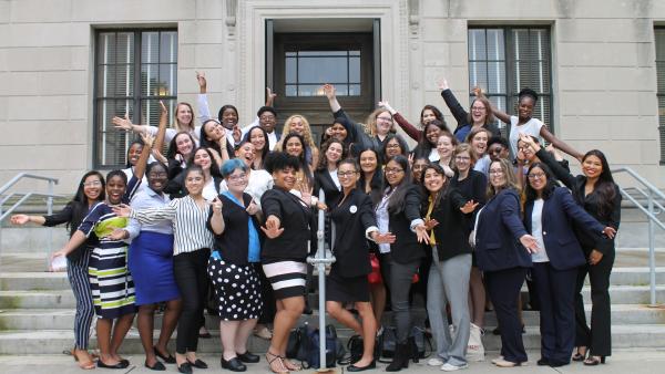 A large group of college age women smile on concrete steps in business attire with their arms outstretched