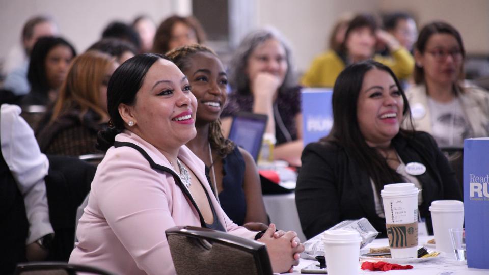 Women of different ages and races smile as they sit at tables in a conference room