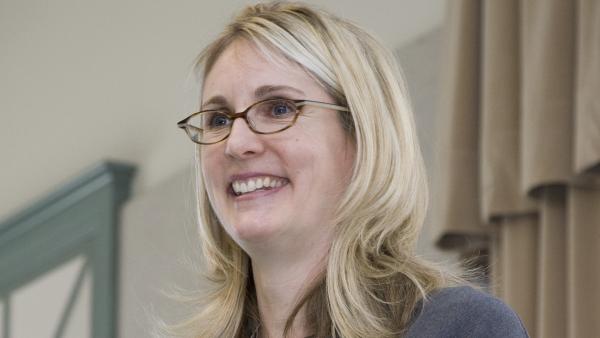 Woman with blond hair and glasses wearing a black shirt and gray jacket.