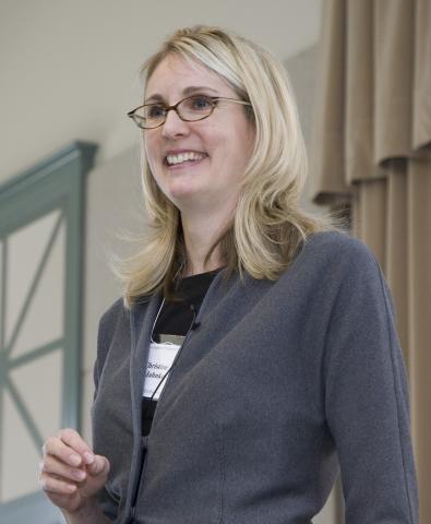 Woman with blond hair and glasses wearing a black shirt and gray jacket.