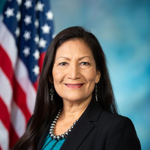 Deb Haaland smiling in front of American flag in black suit jacket