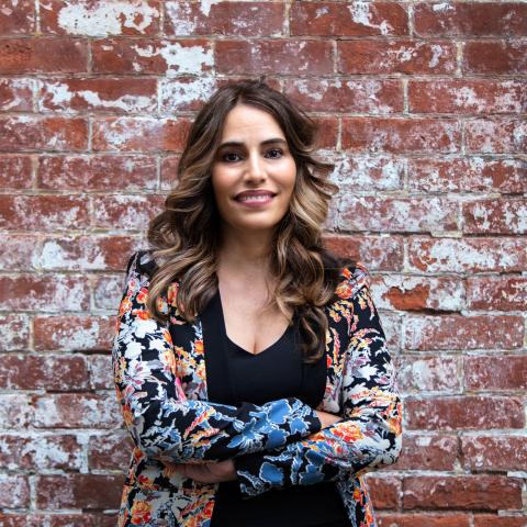 Head shot Senator Teresa Ruiz wearing a multi-color blazer and black shirt, standing with arms crosses against a brick background