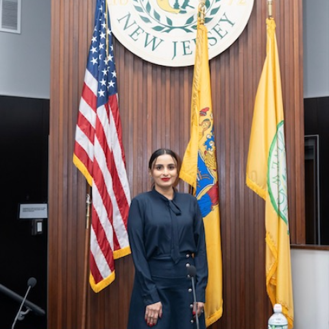 Fozia Janjua poses in all black in front of the Mount Laurel crest, and the flags of the United States and New Jersey