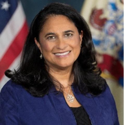 Kellie Doucette seated in front of the flags of the United States and New Jersey. She is wearing a dark blue blazer and black top and has dark brown hair