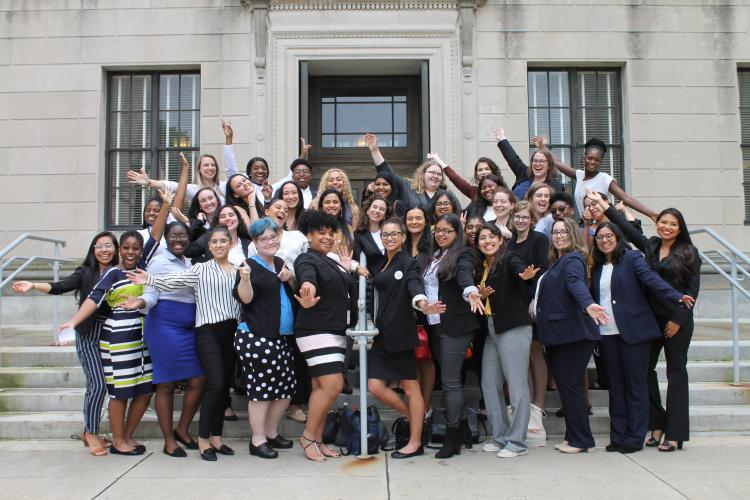 A large group of college age women smile on concrete steps in business attire with their arms outstretched