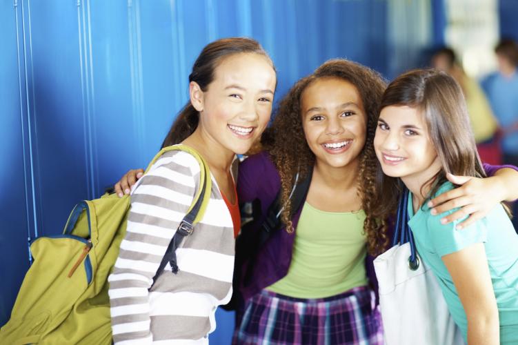 Three school age girls wearing backpacks and school bags smile with arms around one another in front of lockers