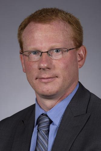 David Andersen in a dark gray suit, blue shirt and tie in front of a gray background