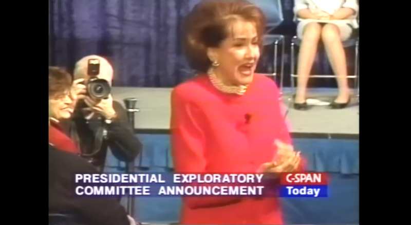 Image of press conference with Elizabeth Dole in red suit in front of stage, photographer taking her picture at right