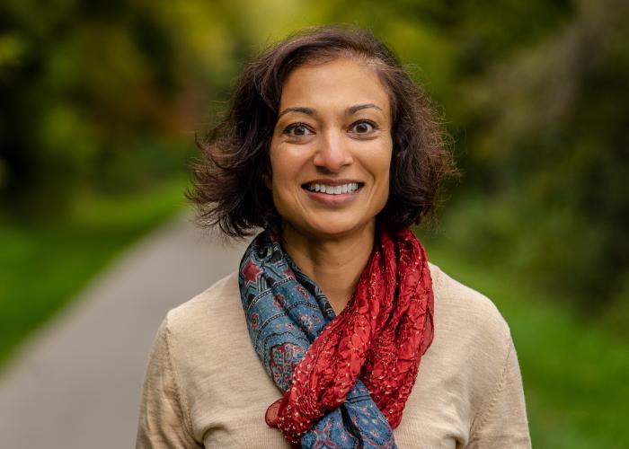 Paru Shah in a beige shirt with a blue and red scarf in front of a blurred outdoor background