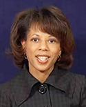 Melody C. Barnes posing in front of dark blue background wearing a black striped jacket with large lapels and big buttons