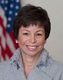 Valerie B. Jarrett posing in front of white background with American flag wearing a silver jacket