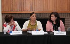 Three women sitting at a panel table, speaker at the center is talking while other participants look at her. 