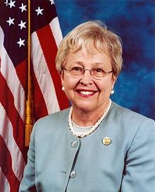 Representative Nancy Johnson wearing green suit with American flag behind her