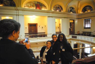 Three young women posing while another takes a photo on a cellphone.