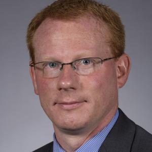 David Andersen in a dark gray suit, blue shirt and tie in front of a gray background