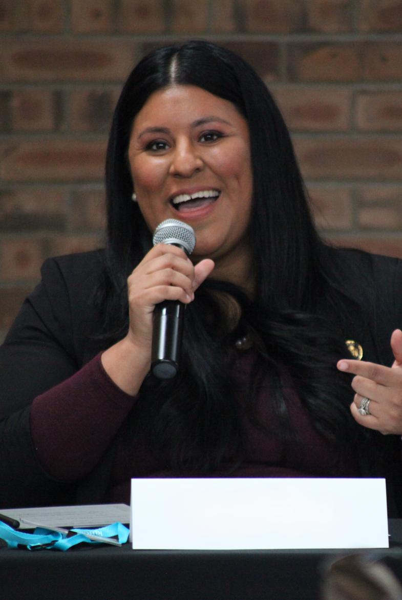 Woman with long dark hair, wearing a black blazer jacket, holding a microphone 