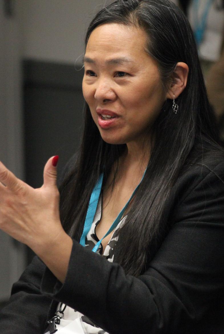 Woman with long dark hair, wearing a black blazer, holding her hand up