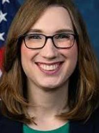 Sarah McBride, wearing black glasses with brown shoulder-length hair, a black suit jacket, standing in front of an American flag.