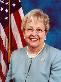 Representative Nancy Johnson wearing green suit with American flag behind her